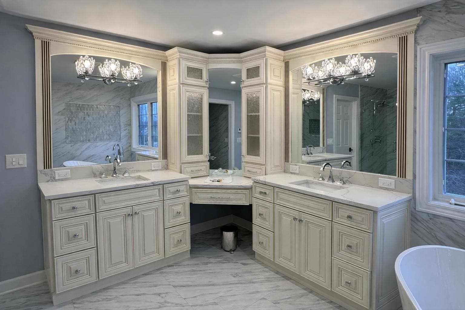Off-white raised-panel double vanity with two countertop storage towers, a glass-front corner cabinet, quartz countertops, twin arched mirrors with crystal light fixtures and a seated makeup station between the two sink runs in a Long Grove, IL primary bathroom